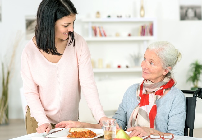 Femme souriante qui donne un plateau repas à une dame en fauteuil