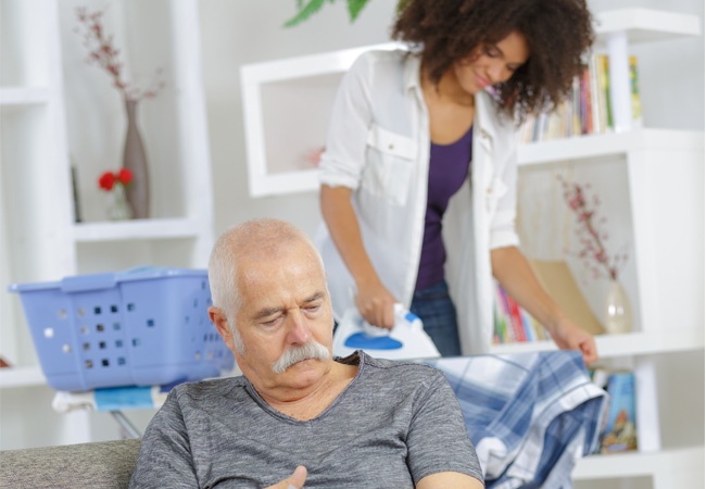 Une jeune femme souriant repasse le linge d'un monsieur en situation de handicap qui lit