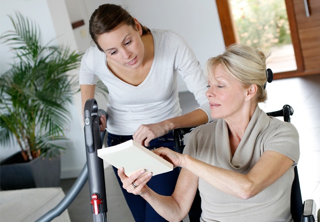 Une femme souriante passe l'aspirateur chez une dame en fauteuil roulant